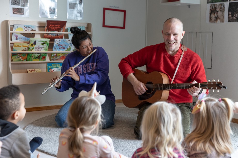 En mann og en dame sitter på gulvet og spiller instrumenter foran en gruppe barn